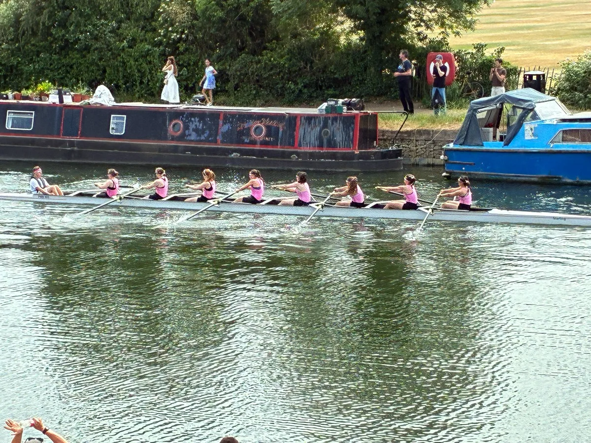 Pembroke College women's first VIII rowing past moored college barges on the Isis during Summer Eights 2026, in their pink kit
